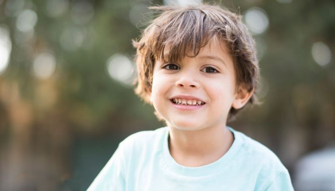 Enfant souriant à l'extérieur, vêtu d'une chemise bleu clair, sur fond flou.