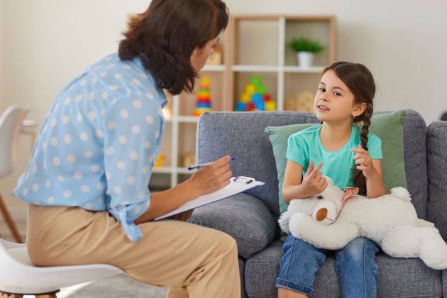 Une thérapeute discute avec un enfant assis sur un canapé dans un salon lumineux, un carnet à la main, l'enfant tenant une peluche.