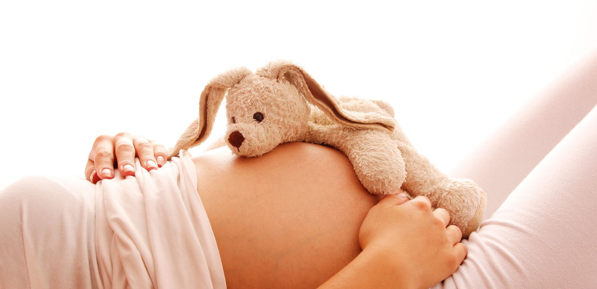 Un ventre de femme enceinte, des mains posées dessus, surmonté d'un petit lapin en peluche baigné d'une douce lumière.