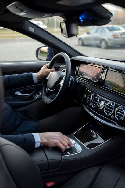 Le conducteur, à l'intérieur d'une voiture de luxe, les mains sur le volant et le sélecteur de vitesse, aperçoit le tableau de bord et la rue.
