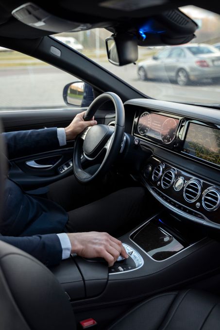Le conducteur, à l'intérieur d'une voiture de luxe, les mains sur le volant et le levier de vitesse, avec un écran tactile et des aérations visibles sur le tableau de bord.