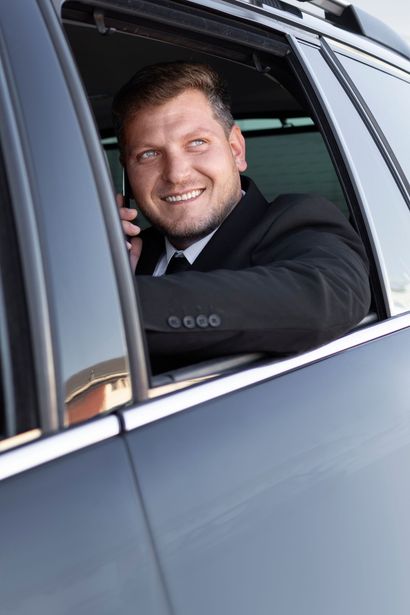 Un chauffeur souriant en costume noir se penche par la fenêtre d'une voiture sombre.