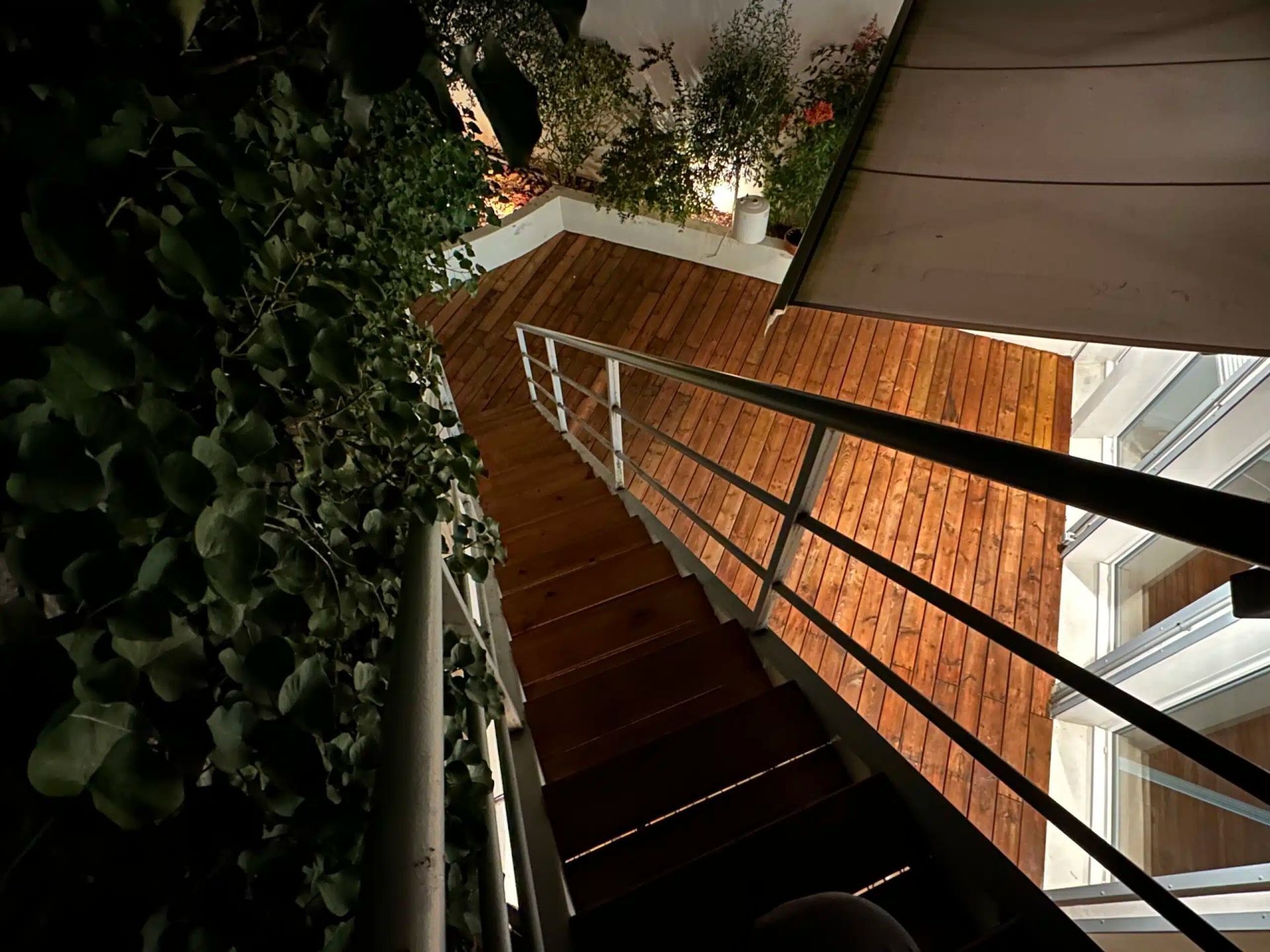 Escalier intérieur avec rampes en métal, longeant un mur de briques et de luxuriantes plantes suspendues.