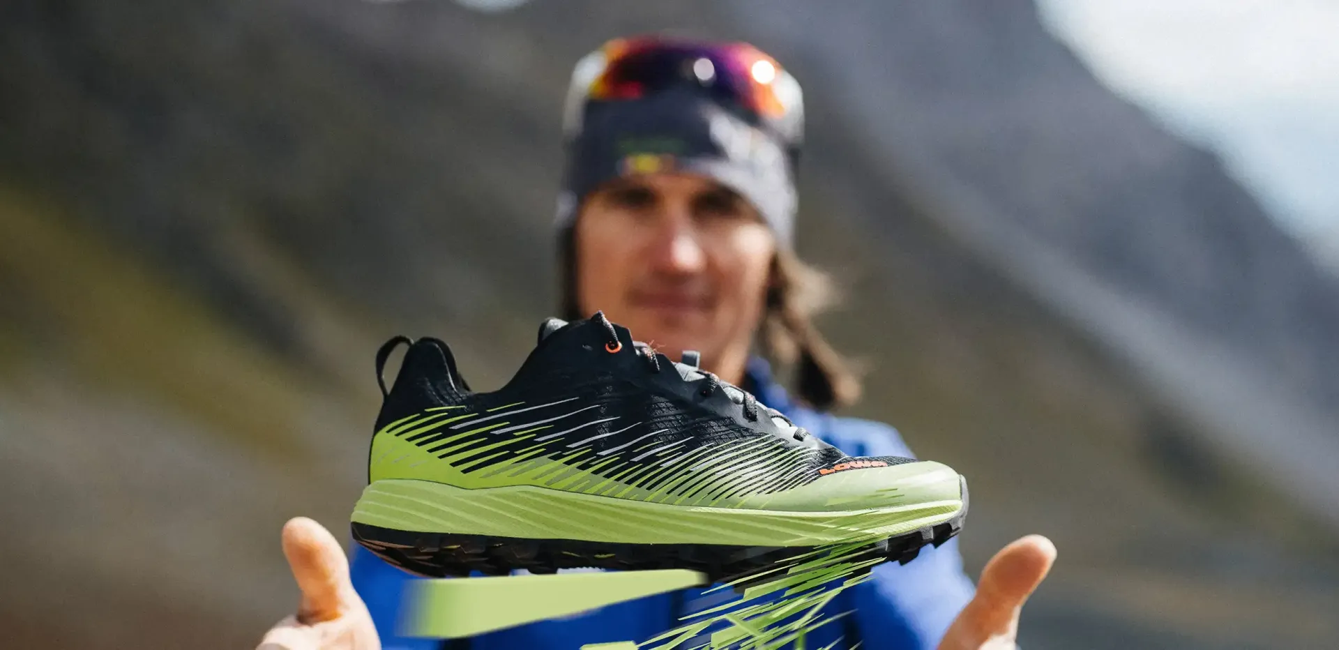 Ein schwarz-neongrüner Laufschuh stand im Freien, im Hintergrund waren eine Person und Berge verschwommen zu erkennen.
