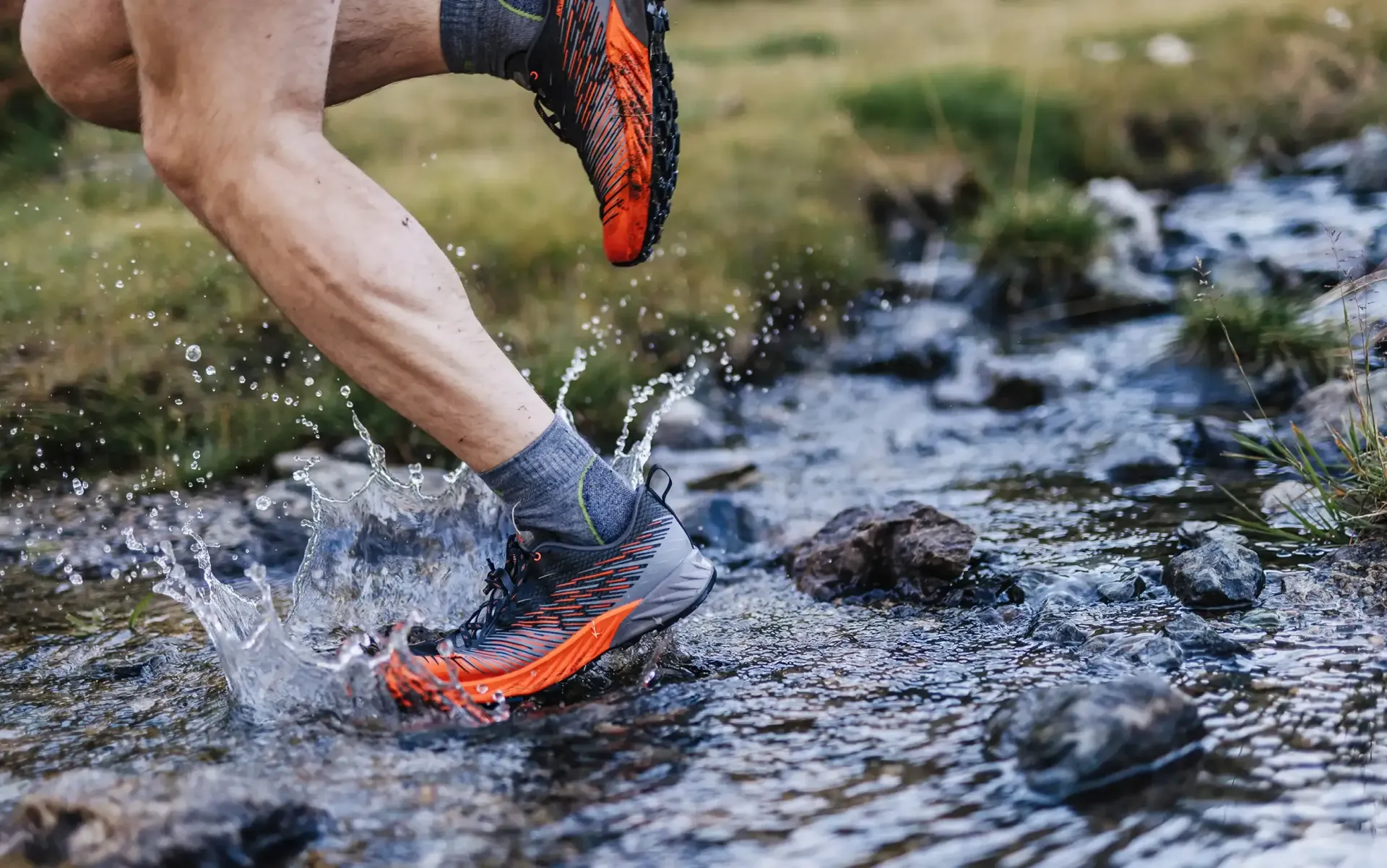 Läuferbeine waten in orange-schwarzen Trailrunning-Schuhen durch einen flachen, schlammigen Bach.