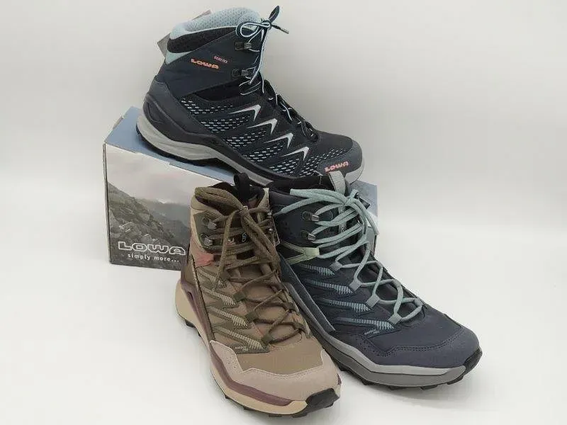 Drei Wanderschuhe in Schwarz, Blau und Beige sind auf einem Karton vor einem weißen Hintergrund gestapelt.