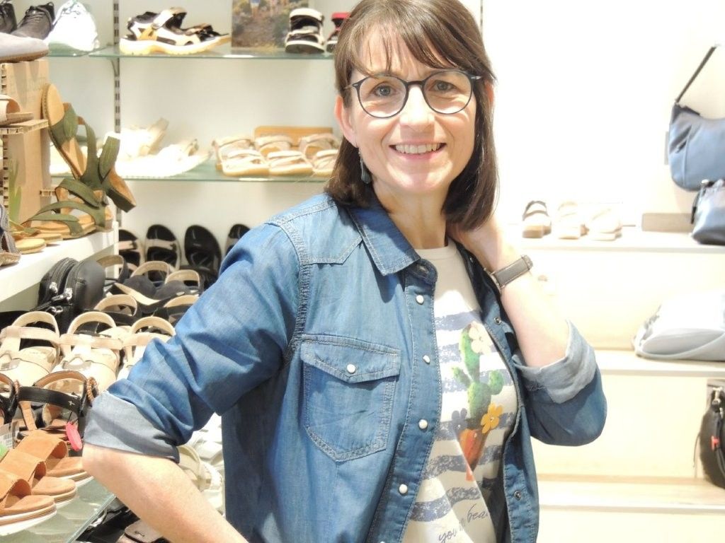 Eine lächelnde Frau mit Brille posiert in einem Schuhgeschäft. Sie trägt ein Jeanshemd und ein geblümtes Top.