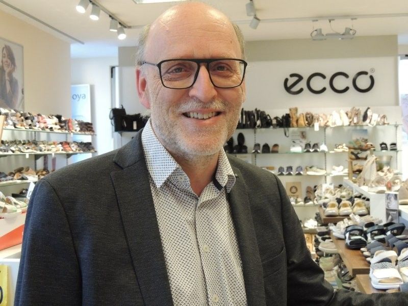 Ein lächelnder Mann mit Brille steht in einem Schuhgeschäft mit ECCO-Logo und Schuhregalen im Hintergrund.