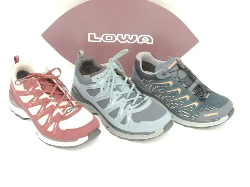 Drei Lowa-Trailrunningschuhe in Rosa, Hellgrau und Dunkelgrau sind vor einem LOWA-Logo angeordnet.