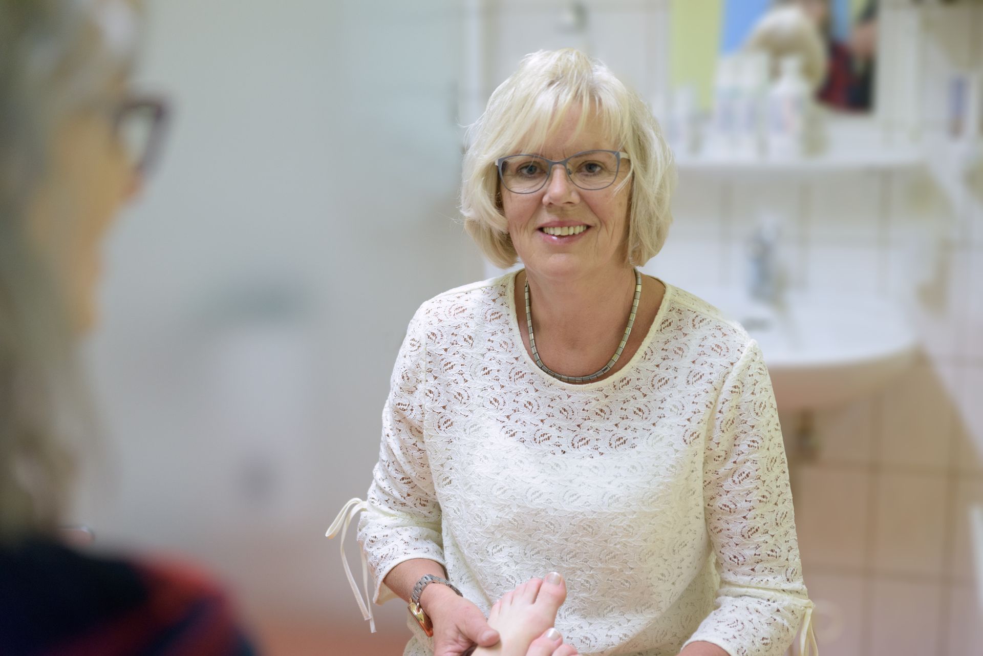 Eine lächelnde Frau in einem weißen Pullover steht in einer hellen Klinik- oder Büroumgebung.