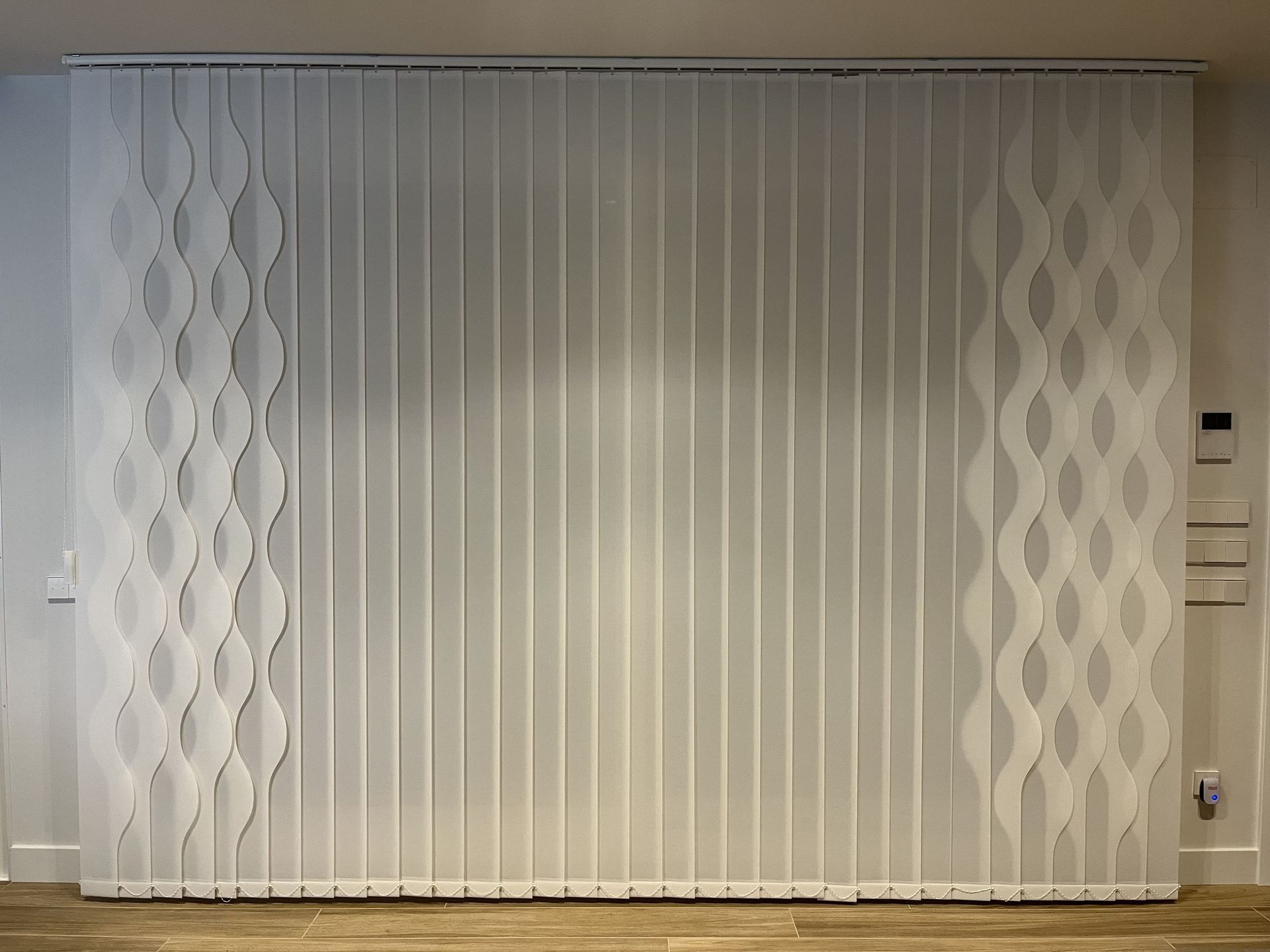 Una persiana vertical blanca con un patrón está colgada en una pared.