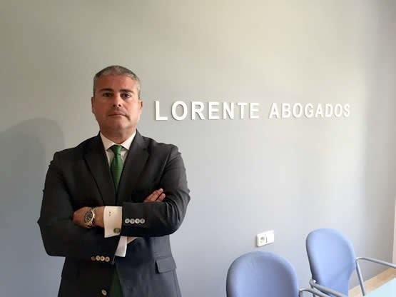 Mujer con cabello castaño rizado, gafas y un blazer, sosteniendo una computadora portátil, sonriendo.