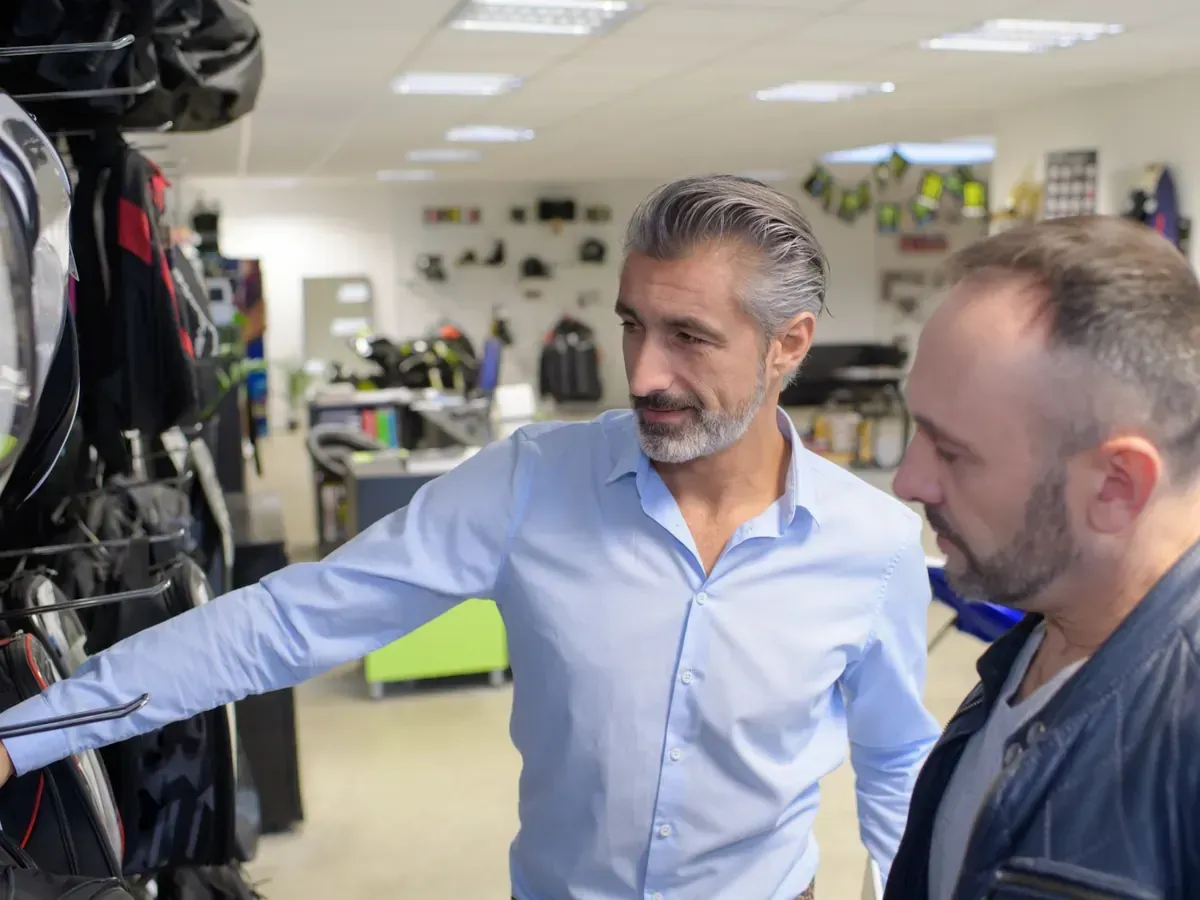 Dos hombres en una tienda, uno de ellos señalando un casco. Estantes con equipo al fondo.