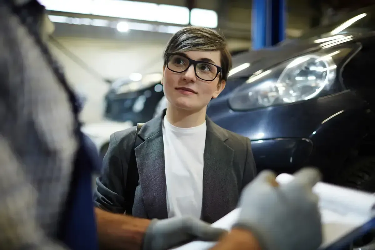 Mujer con gafas hablando con un mecánico en un taller mecánico; coche al fondo.