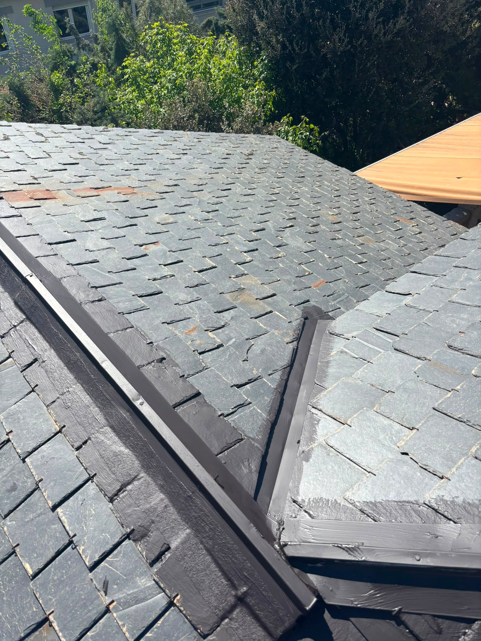 Techo de tejas grises con una ventilación de cumbrera dañada y árboles al fondo.
