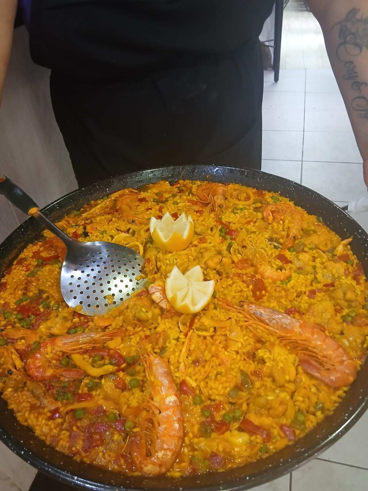 Gran paellera con arroz al azafrán, gambas y rodajas de limón, con una cuchara para servir encima.