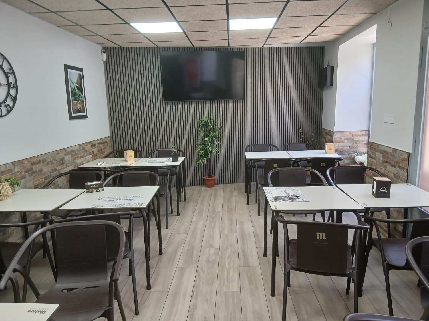 Comedor vacío de un restaurante con mesas, sillas, suelo de madera, televisor de pared y una planta en maceta al fondo.