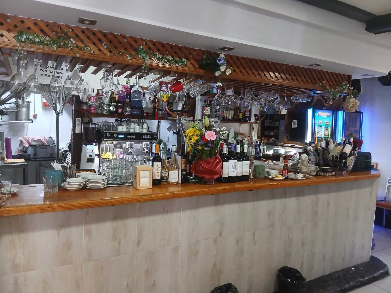 Barra de bar en una acogedora cafetería con vasos colgantes, estantes, máquinas de café y decoración floral.