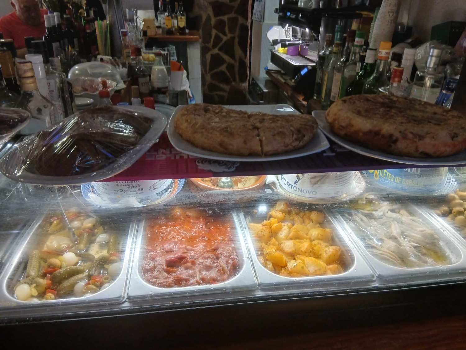 Mostrador de charcutería con bandejas de comida preparada y dos pizzas grandes bajo una vitrina de cristal.