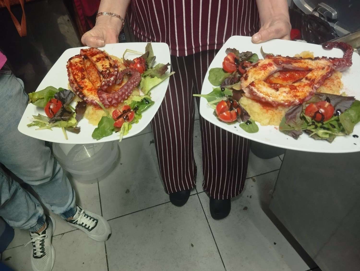 Dos personas sosteniendo platos de pollo asado con verduras sobre un suelo de baldosas en la cocina.