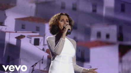Mujer con vestido blanco cantando en el escenario; edificios blancos en el fondo.