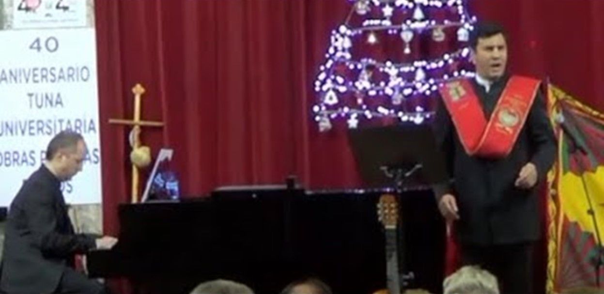 Un hombre canta al piano acompañado de un pianista. Se ven cortinas rojas y un árbol de Navidad.