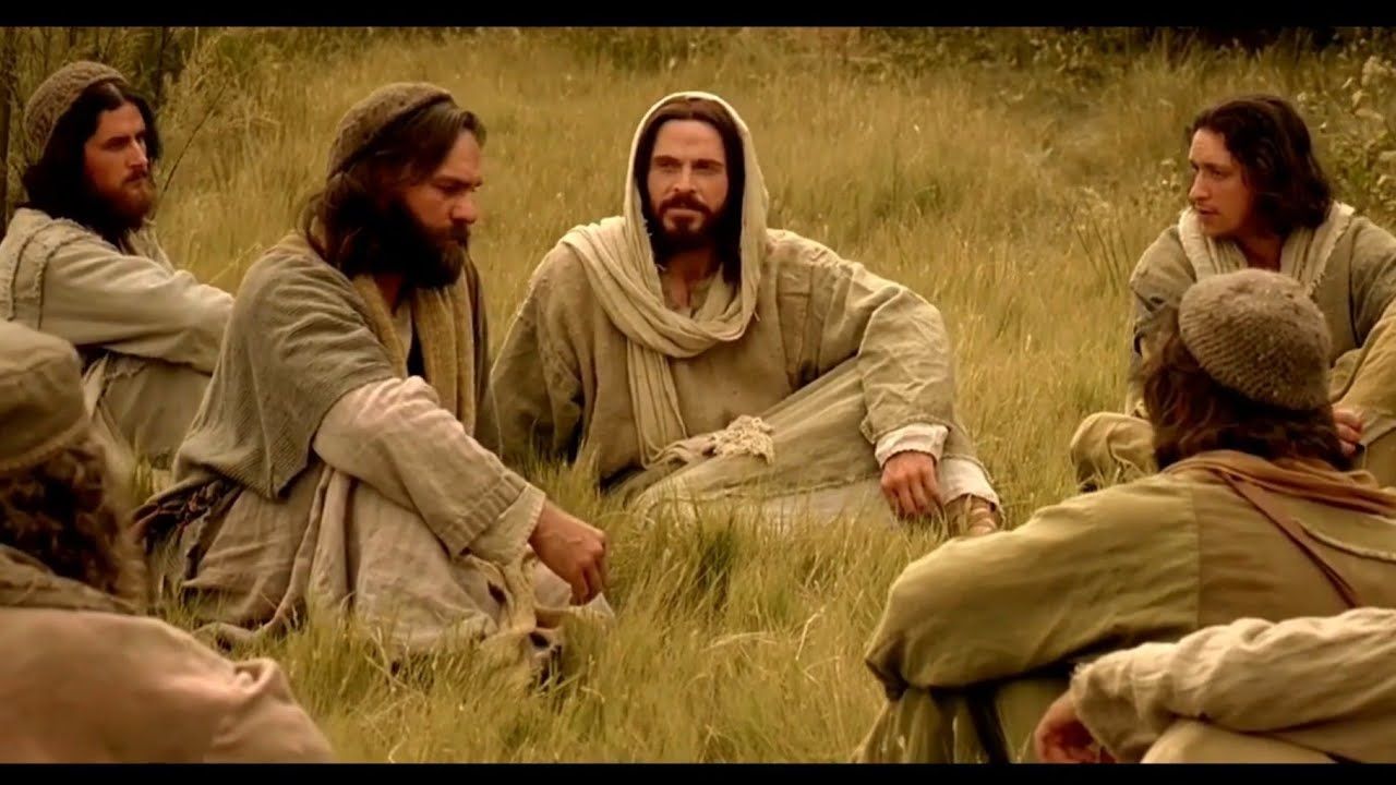 Jesús y sus discípulos conversan sentados en un campo de hierba. Luz tenue, tonos neutros, expresiones contemplativas.