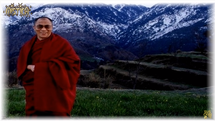 El Dalai Lama, vestido de rojo, sonriendo, de pie frente a montañas cubiertas de nieve, campos verdes y colinas con terrazas.
