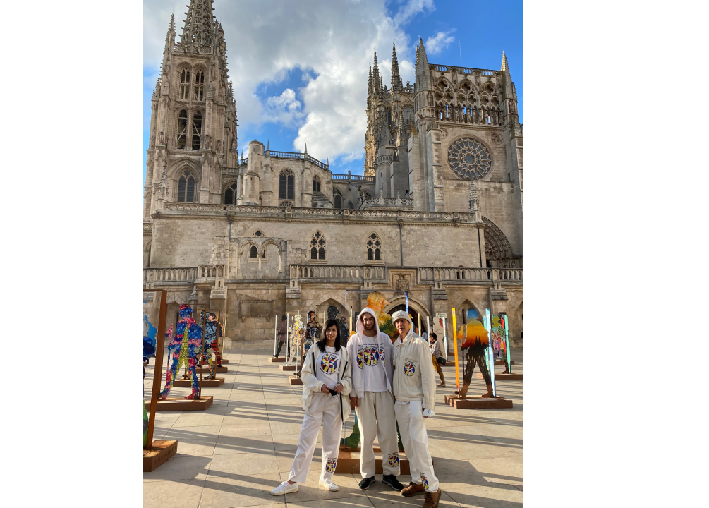 Tres personas posan frente a la Catedral de Burgos en España, con coloridas exhibiciones de arte cerca; visten trajes blancos.