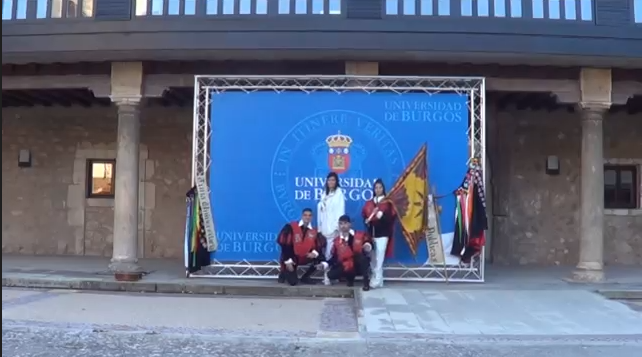 Un grupo de personas vestidas con atuendo académico posan frente a un telón universitario; banderas a ambos lados.