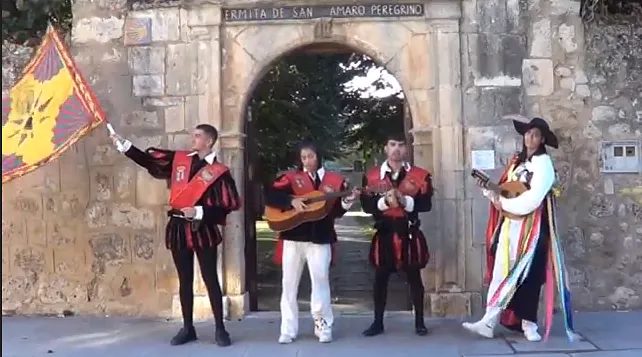 Cuatro músicos disfrazados frente a un arco de piedra. Uno sostiene una bandera, mientras los demás tocan instrumentos de cuerda.