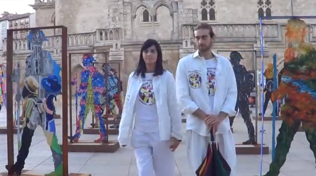 Dos personas vestidas de blanco caminan junto a figuras recortadas de colores en una plaza.