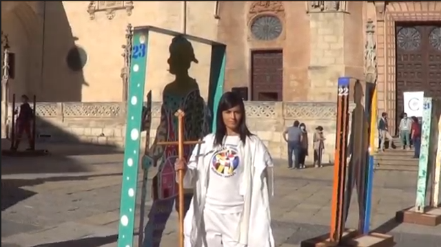 Una mujer vestida de blanco se sitúa delante de una obra de arte que presenta sombras, en una plaza.