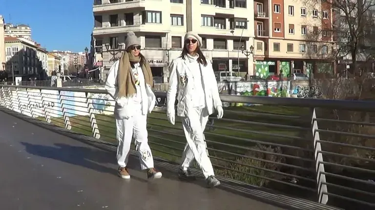 Dos personas vestidas de blanco caminando por un puente en una ciudad, en un día soleado.
