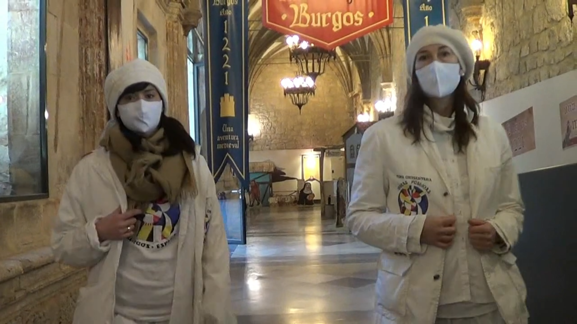 Dos mujeres con batas blancas y mascarillas en un salón con una pancarta de 