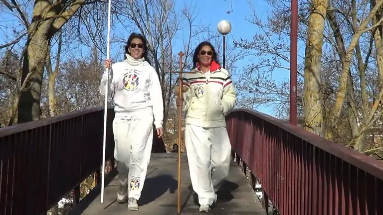 Dos personas, vestidas de blanco y sosteniendo bastones, caminan por un puente. Día soleado.