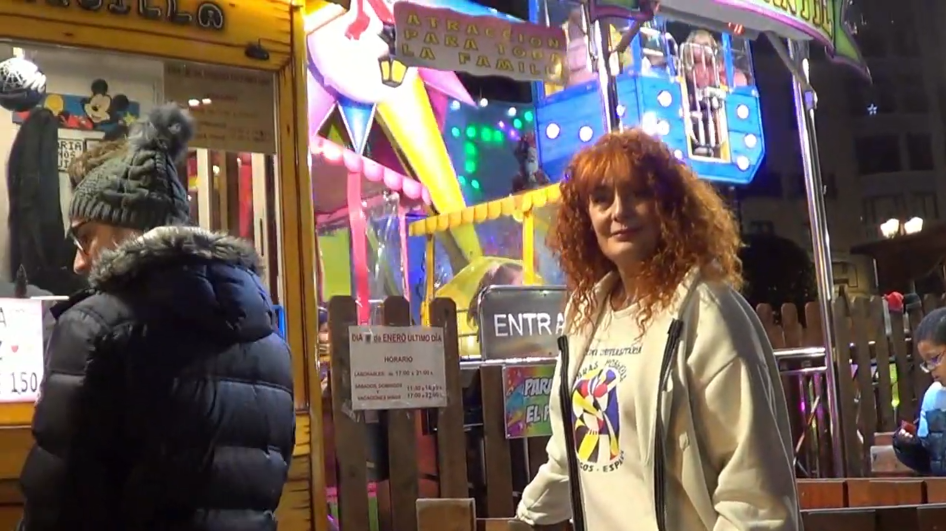 Una mujer pelirroja sonríe junto a una atracción de feria. Cerca hay una persona con chaqueta negra.