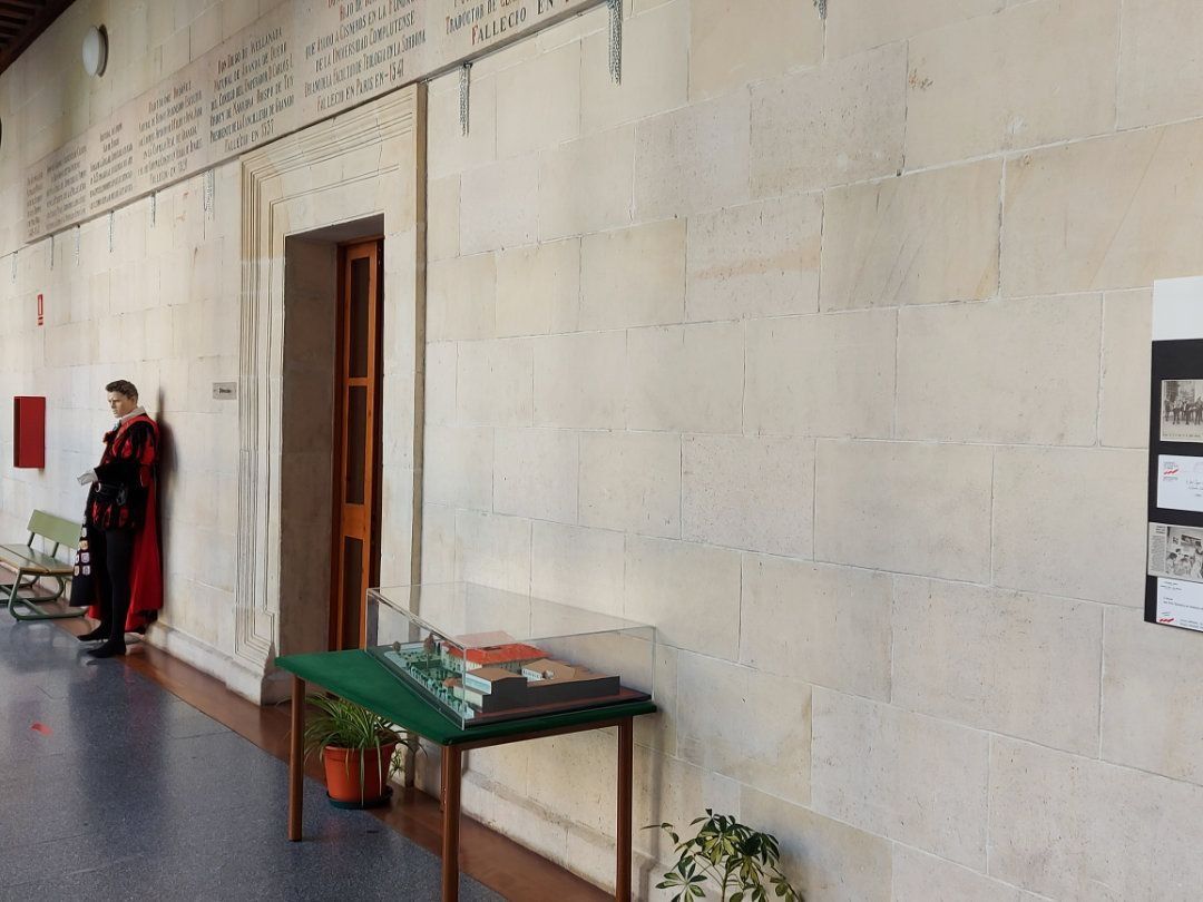 Una persona vestida formalmente se apoya contra una pared de piedra clara en un pasillo, cerca de una puerta y una mesa de exhibición.