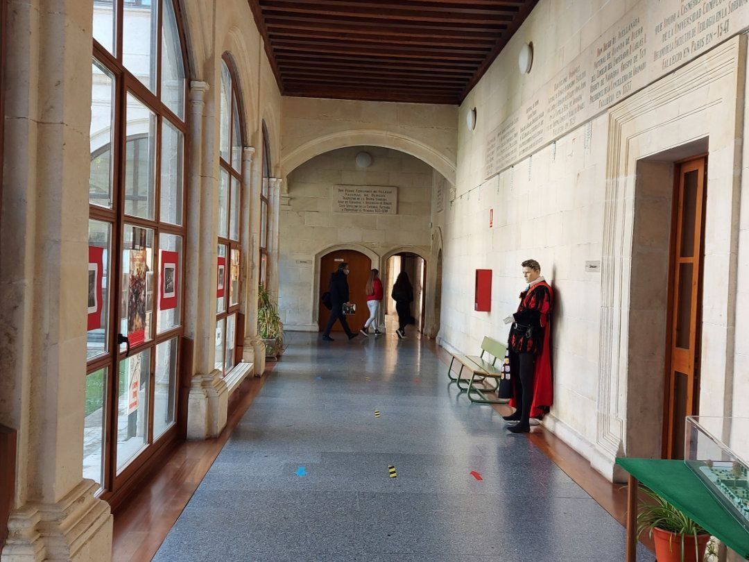 Pasillo con puertas arqueadas, ventanas y una figura con una capa roja apoyada contra una pared.