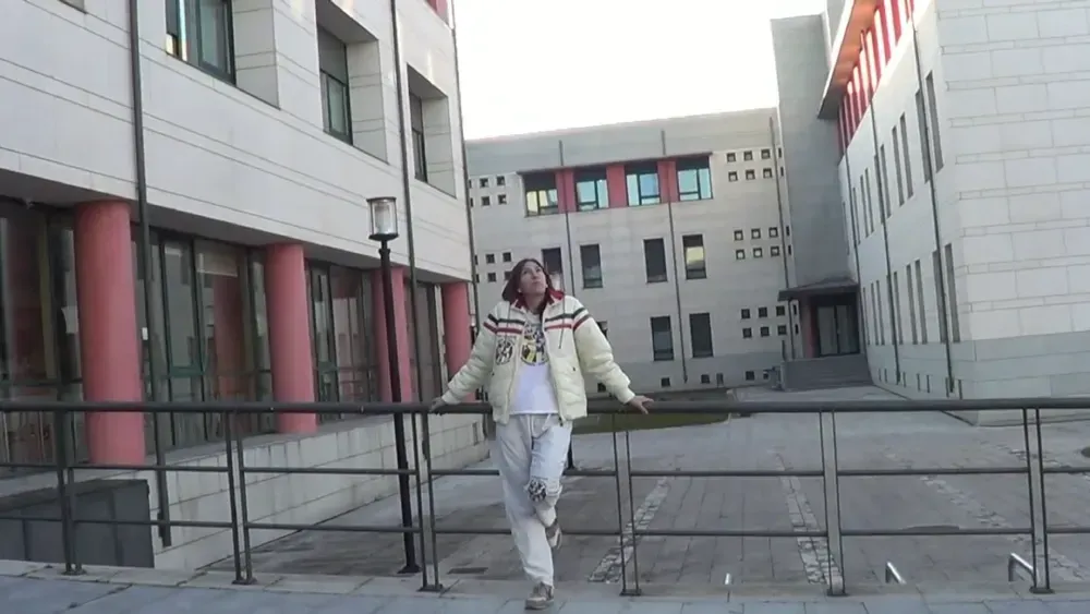 Persona parada en un puente frente a edificios; vistiendo una chaqueta blanca y pantalones gris claro; durante el día.