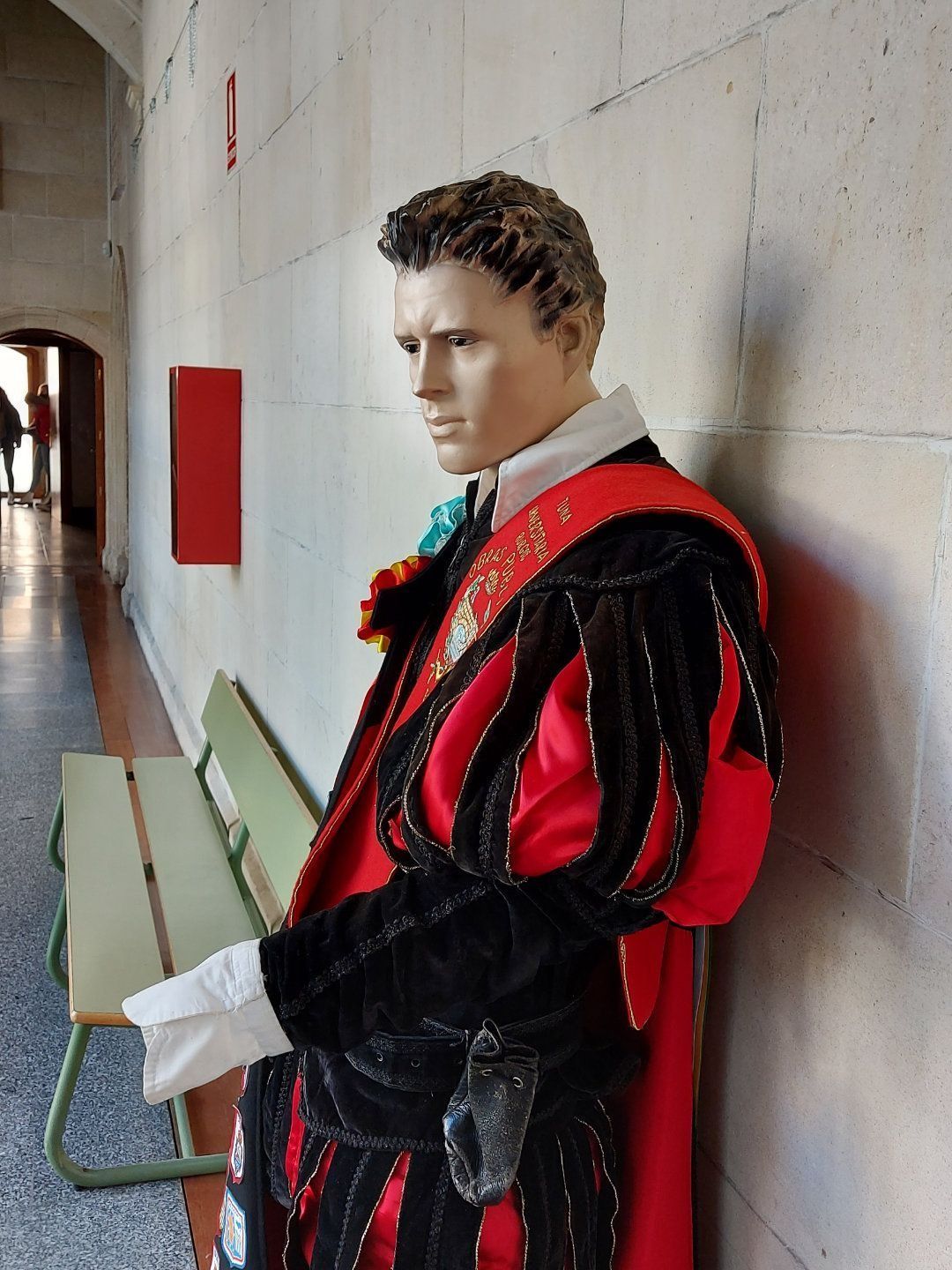 Un maniquí con ropa roja y negra de estilo renacentista se apoya contra una pared en el pasillo de un edificio.