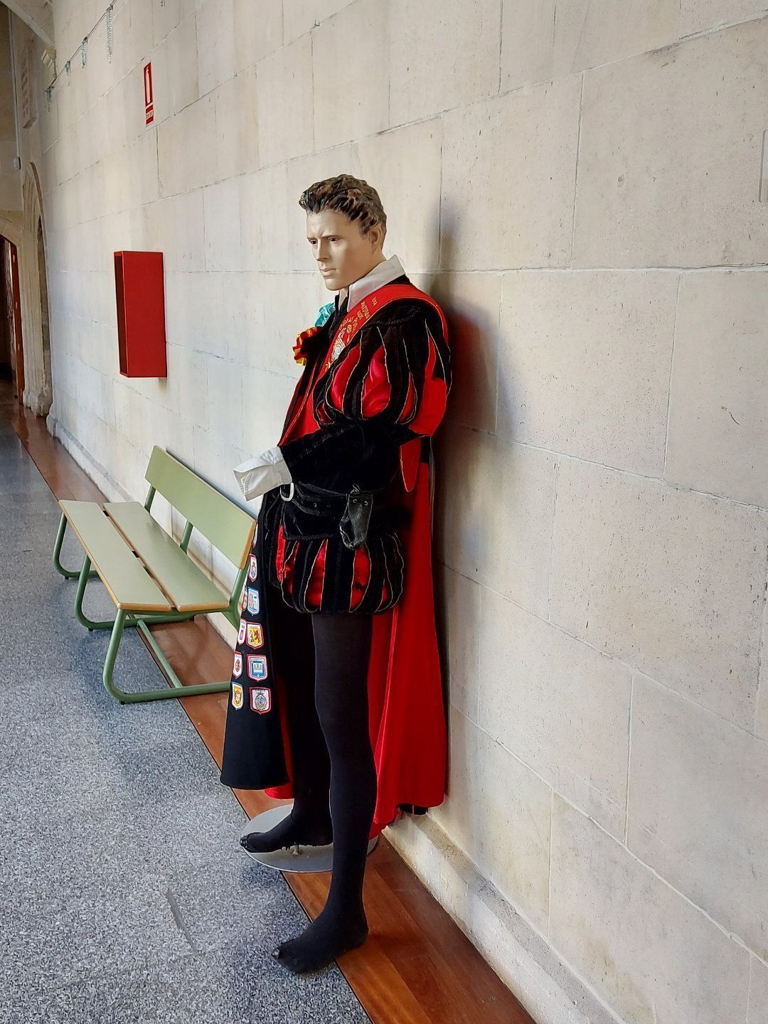 Un maniquí con traje renacentista se apoya en la pared de un pasillo, cerca de un banco.