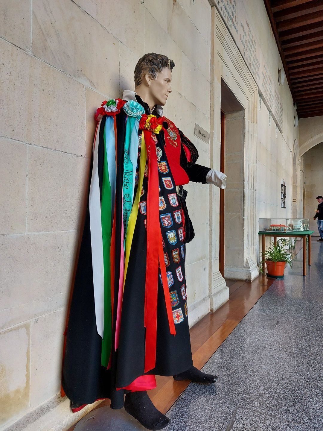 Maniquí con túnica negra y cintas de colores, apoyado contra una pared de piedra en un edificio.