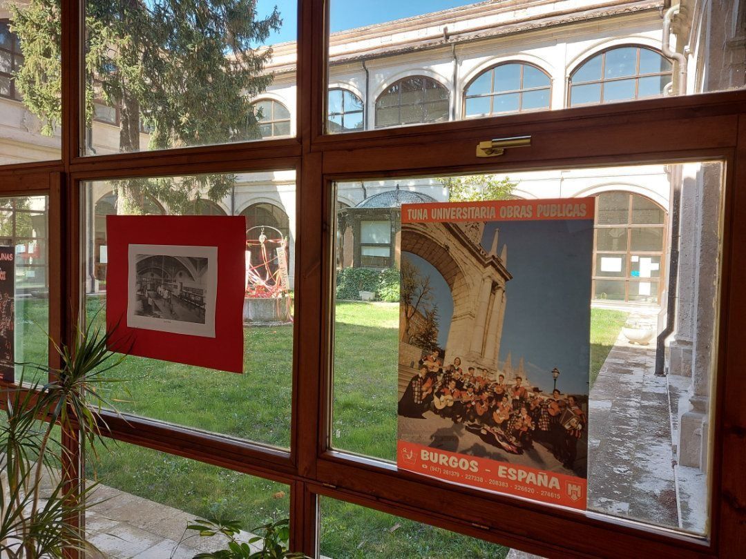 Vista desde la ventana: patio con carteles sobre vidrio. Arte con marco rojo, póster colorido con figuras, césped verde.