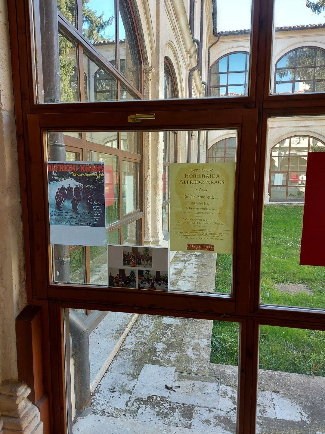 Ventana con marco de madera con carteles en el interior; vista de un patio de piedra con arcos y césped.