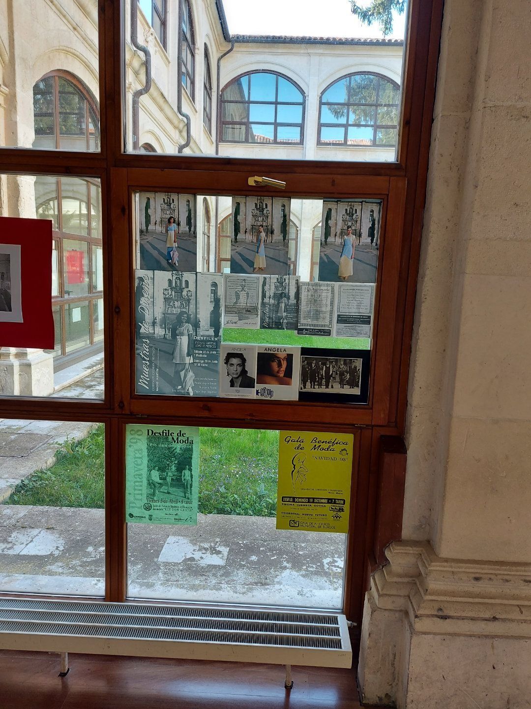 Escaparate con carteles y fotos en el patio de un edificio. Marco marrón, césped verde visible en el exterior.
