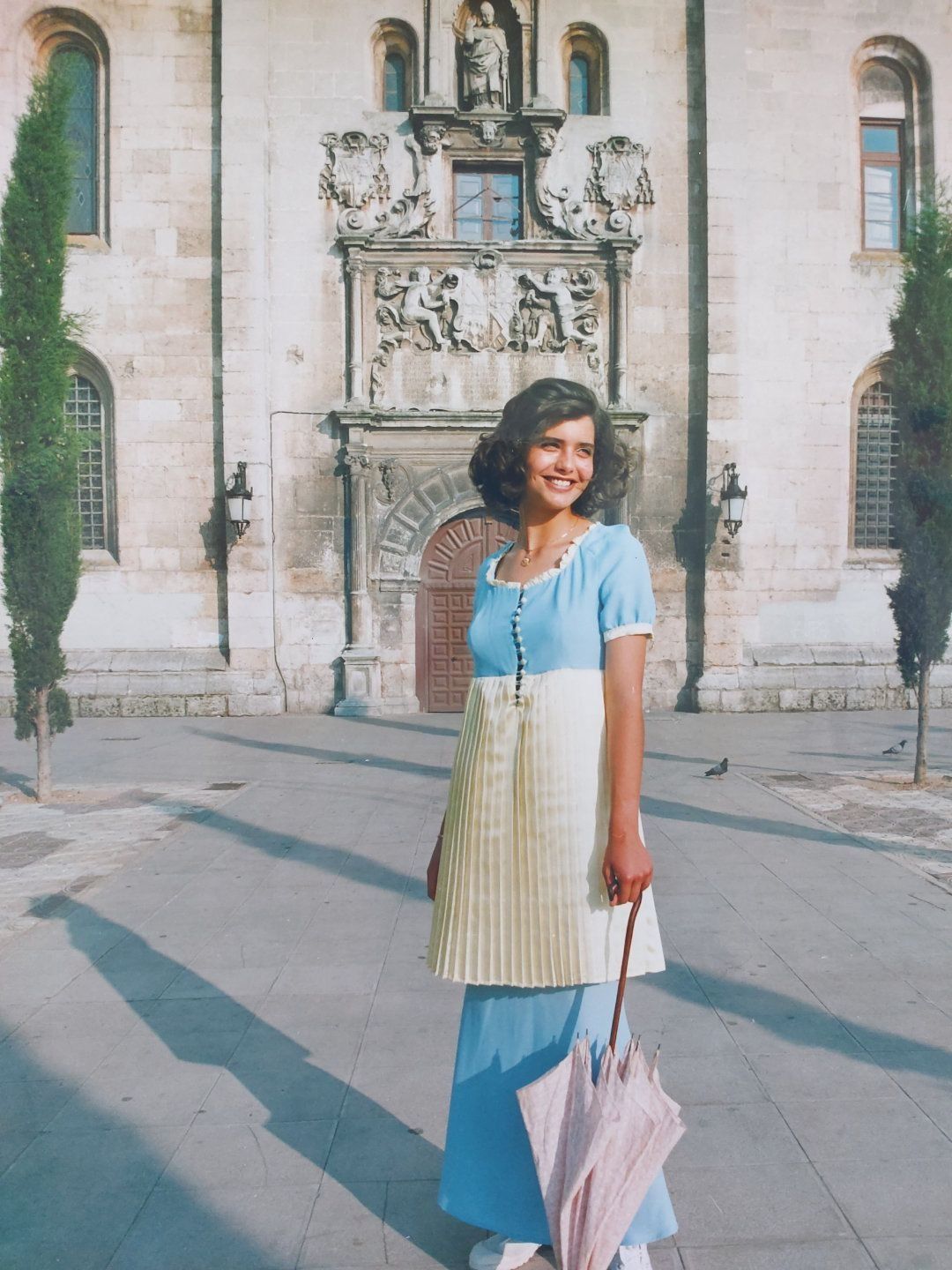 Una mujer con un vestido azul y un paraguas se encuentra frente a un edificio ornamentado bajo la luz del sol.