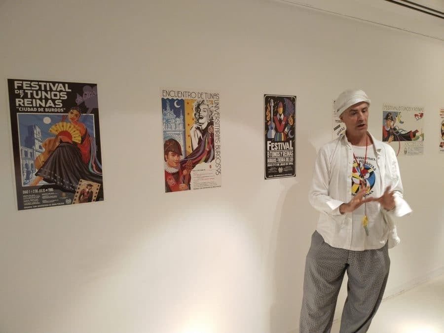 Hombre gesticulando, de pie en una galería con carteles en la pared. Lleva traje blanco y sombrero.