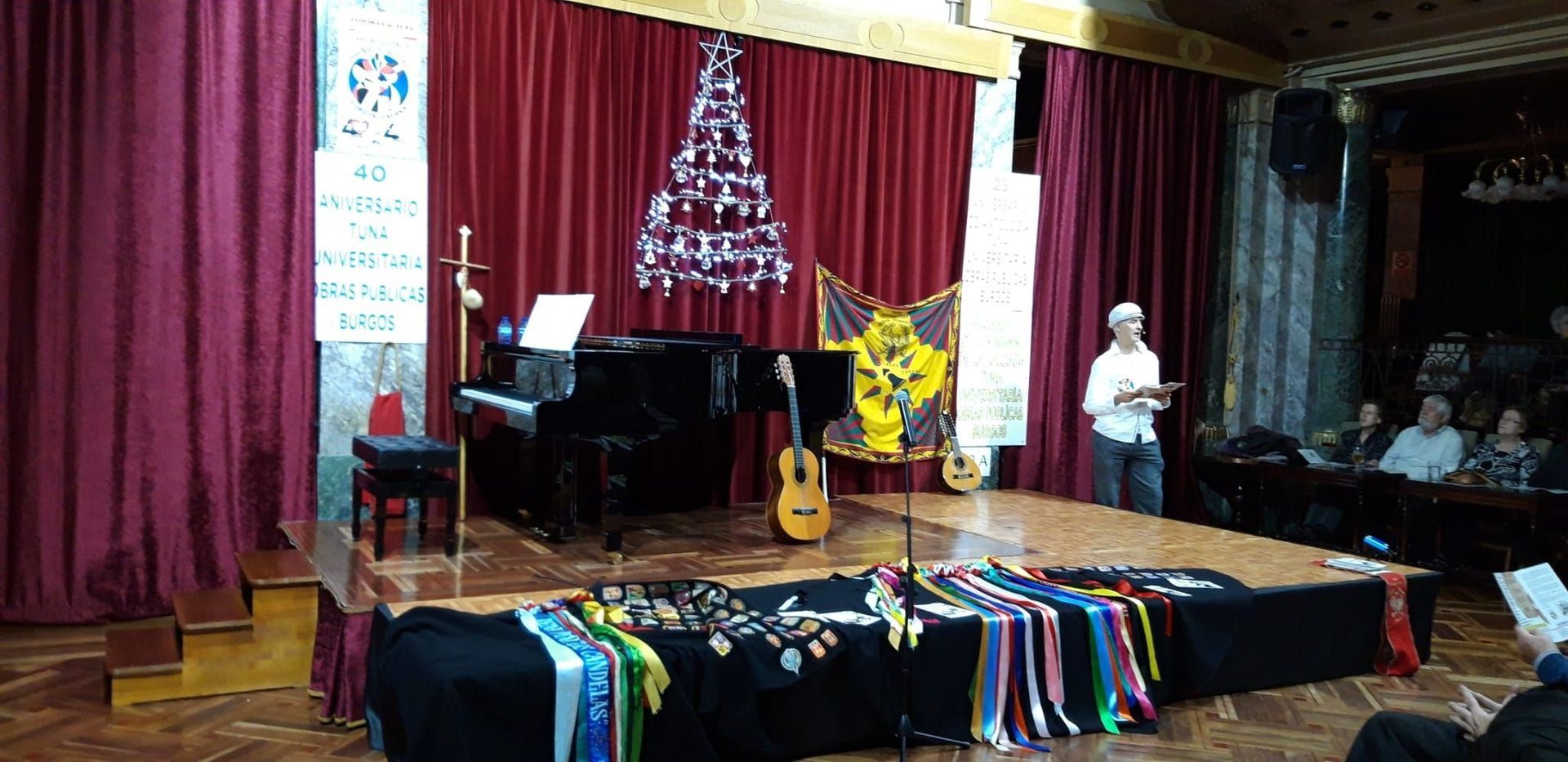 Hombre hablando en el escenario con piano, guitarra y premios frente a una cortina roja. El público está sentado.