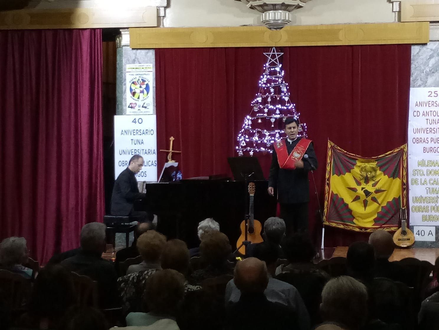 Hombre cantando en el escenario con pianista, público, árbol de Navidad y pancartas en una sala.